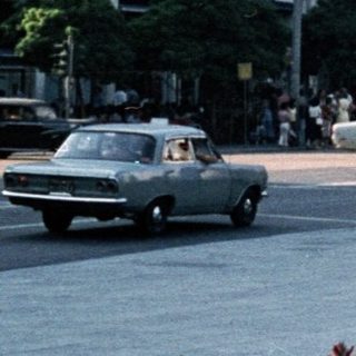 People walking in a city square with vintage cars and floral planters.