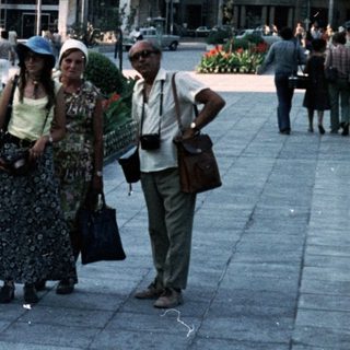 People walking in a city square with vintage cars and floral planters.
