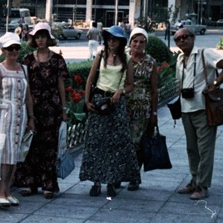 People walking in a city square with vintage cars and floral planters.