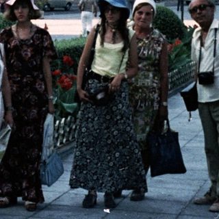 People walking in a city square with vintage cars and floral planters.