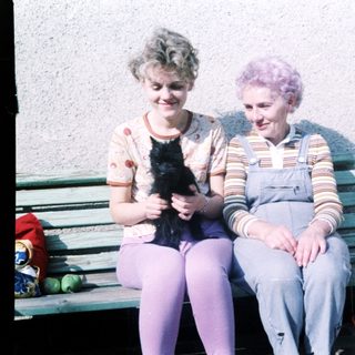 Two women sitting on bench holding black cat.