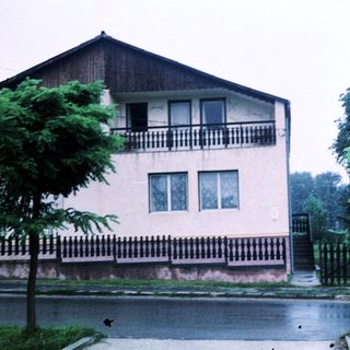 Two-story house with balcony, surrounded by greenery.