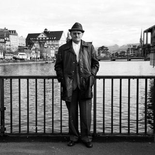 Man in 1960s attire posing by waterfront railing.