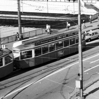 Urban street scene with tram, bridge, and mid-century buildings.