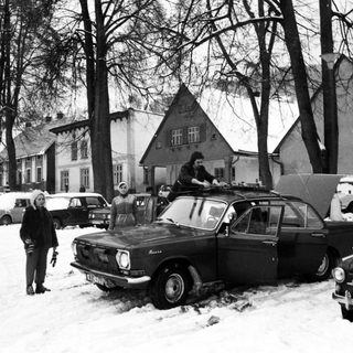 Snowy street scene with vintage cars and people clearing snow.