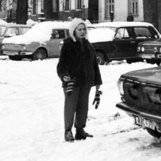 Snowy street scene with vintage cars and people clearing snow.