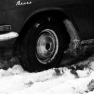 Snowy street scene with vintage cars and people clearing snow.