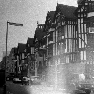 Black-and-white street scene with Tudor-style buildings and vintage cars.