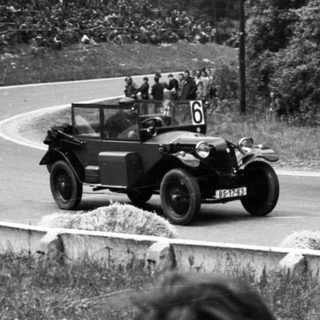 Classic race car driving on winding road with spectators.