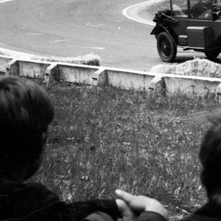 Classic race car driving on winding road with spectators.