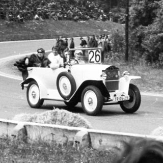 Classic race car driving on winding road with spectators.