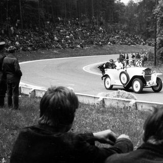 Classic race car driving on winding road with spectators.