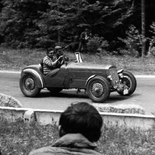 Classic race car driving on public road with spectators.