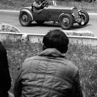 Classic race car driving on public road with spectators.