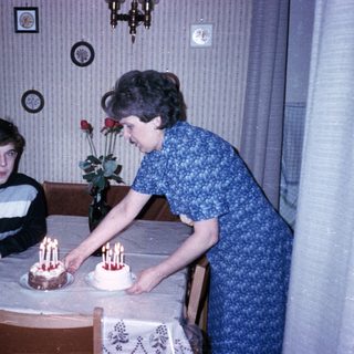 A woman lighting candles on two birthday cakes with a young boy seated.
