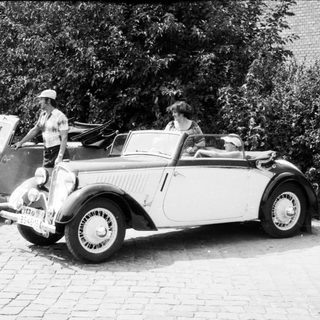 Vintage convertibles parked with people near trees.