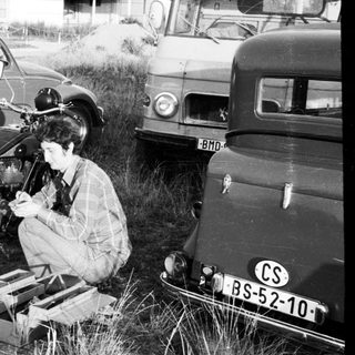 Person repairing motorcycle in grassy field near vintage cars.