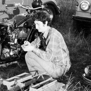 Person repairing motorcycle in grassy field near vintage cars.