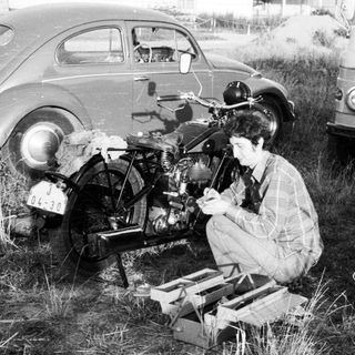 Person repairing motorcycle in grassy field near vintage cars.
