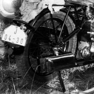 Person repairing motorcycle in grassy field near vintage cars.