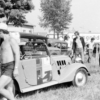Vintage rally car with participants near roadside sign.