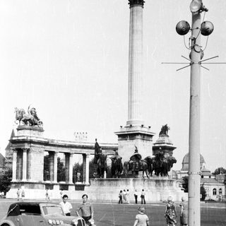 Historic monument with statues, vintage car, pedestrians.