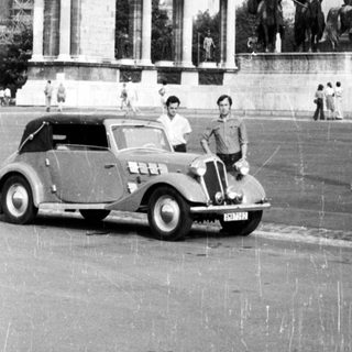 Historic monument with statues, vintage car, pedestrians.
