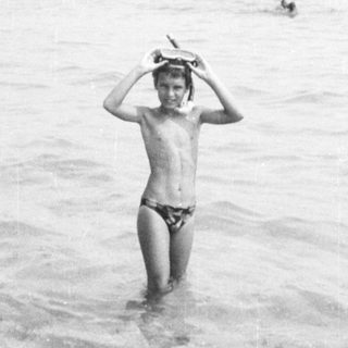 Shirtless boy in shallow saltwater, holding hat.
