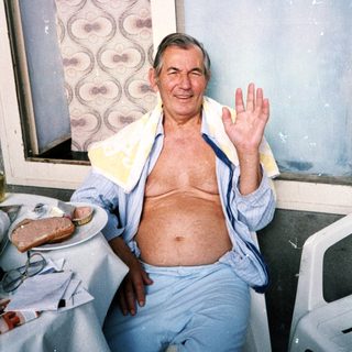 Older man waving shirtless indoors near window.