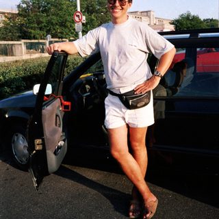 Person standing by open car door wearing shorts, sunglasses, and fanny pack.
