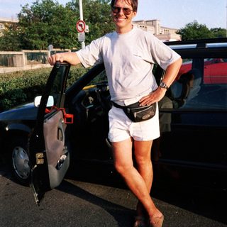 Person standing by open car door wearing shorts, sunglasses, and fanny pack.