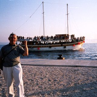 Man poses by pebble shore near crowded ferry boat.