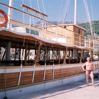 Shirtless man in pink shorts by docked wooden sailboat.
