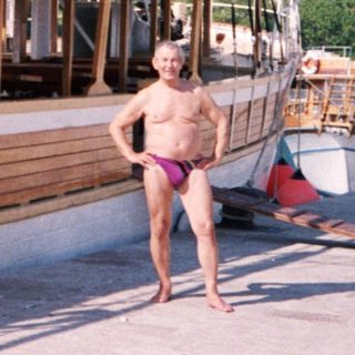 Shirtless man in pink shorts by docked wooden sailboat.