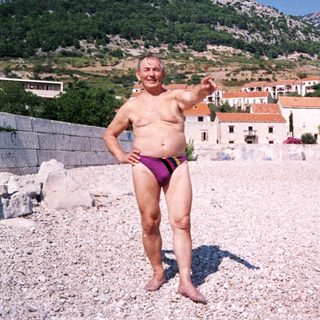 Shirtless man in swim trunks posing on pebble beach.
