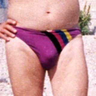 Shirtless man in swim trunks posing on pebble beach.