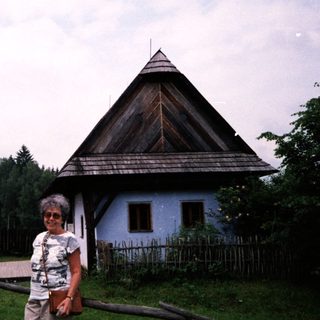 Traditional wooden house with steep roof, woman posing outside.