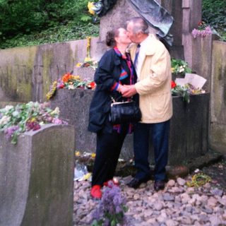 People paying respects at a memorial statue with flowers.