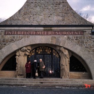 Stone mausoleum entrance with Latin inscription, three people standing beneath archway.