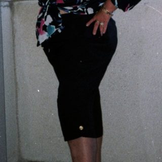 Woman standing in doorway wearing floral top, skirt, and green shoes.