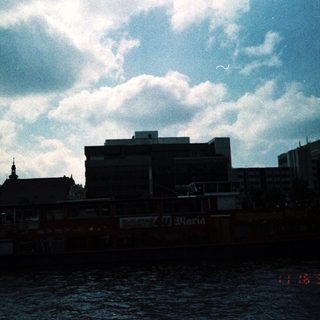 Urban riverside scene with buildings, boats, and dated stamp.