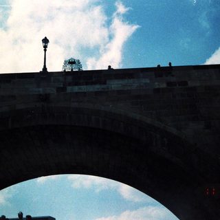 Arch bridge with lamppost and rooftops under cloudy sky.