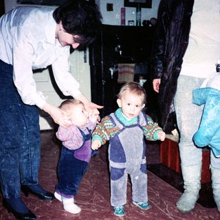 Two adults assist toddlers learning to walk indoors.