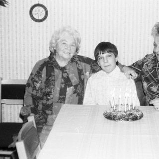 Three generations posing with birthday cake indoors.
