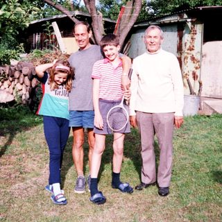Family posing outdoors near rustic cabin with tennis racket.