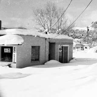 Snow-covered rural street with buried buildings and vehicles.