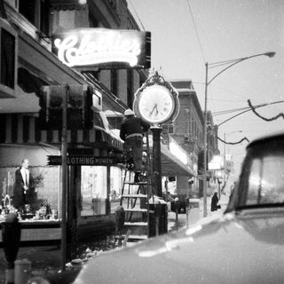 Vintage street scene with clock tower, old car, and storefronts.