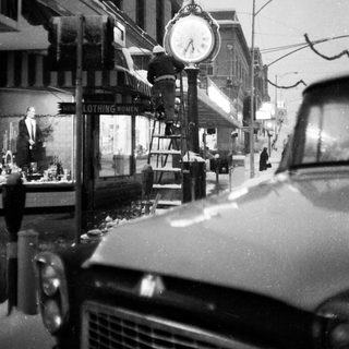 Vintage street scene with clock tower, old car, and storefronts.