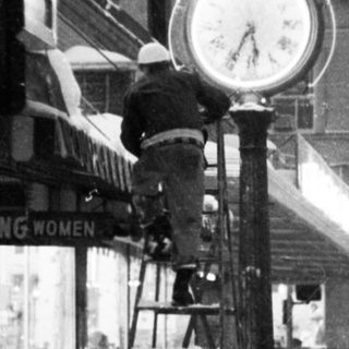 Vintage street scene with clock tower, old car, and storefronts.