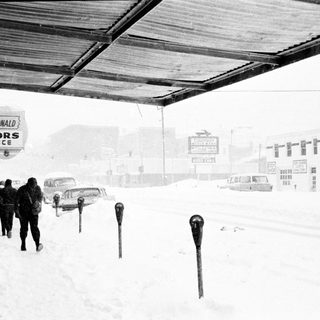 Snowy street scene with pedestrians walking near Curiosity Corner and McDonald Realty signs.